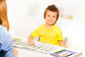 Smiling boy plays in developing game pointing at colorful cards of days and activities on calendar with his parent sitting opposite at the table